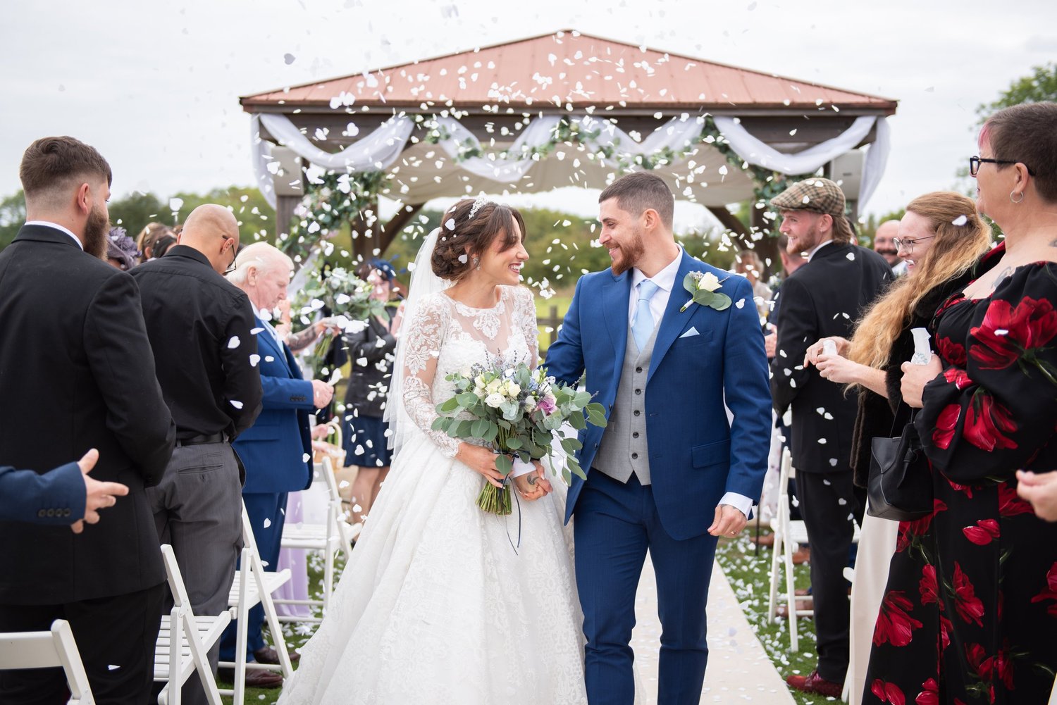 Tom and Bernadette walking back down the aisle, smiling at each other as guests throw confetti